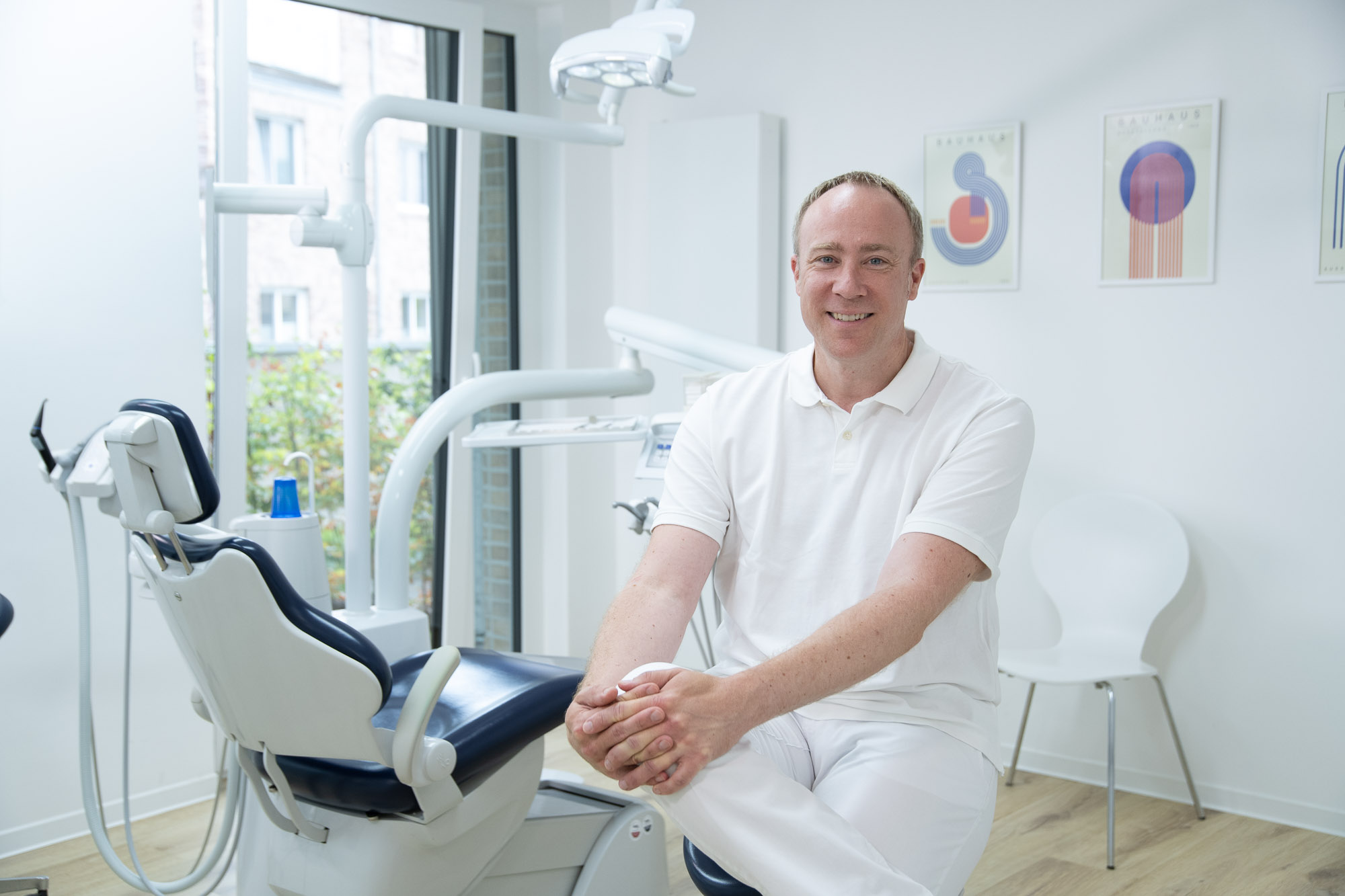 Dr. Christoph Westphal sitzt in weißer Kleidung in seiner modernen Zahnarztbpraxis neben einem Behandlungsstuhl und den Geräten. Der helle Raum hat weiße Wände, helle Holzböden, ein großes Fenster und abstrakte Kunstwerke an der Wand.