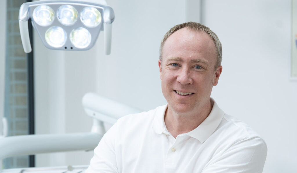 Dr. Christoph Westphal lächelt in einem weißen Hemd in einem hell beleuchteten Behandlungszimmer., Über ihm hängt eine Untersuchungslampe, und im Hintergrund sind zahnärztliche Geräte zu sehen.