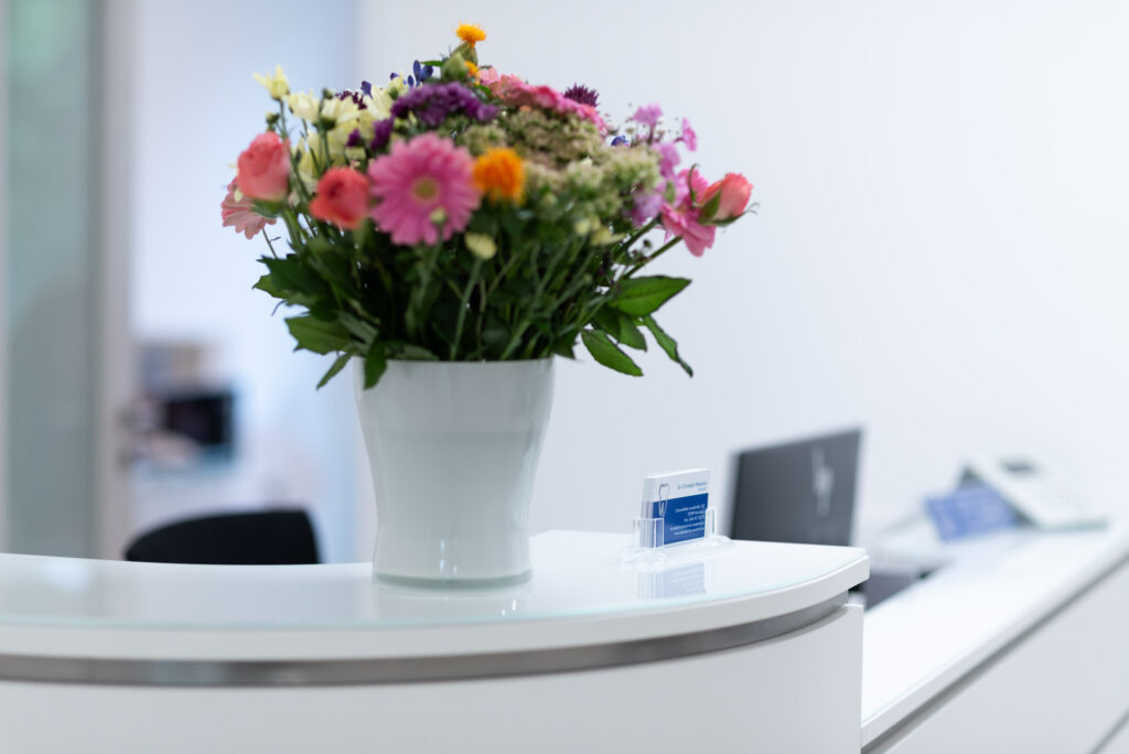 Ein bunter Blumenstrauß in einer weißen Vase steht auf einem modernen, geschwungenen weißen Empfangstresen, im Hintergrund sind verschwommene Bürogeräte zu sehen.